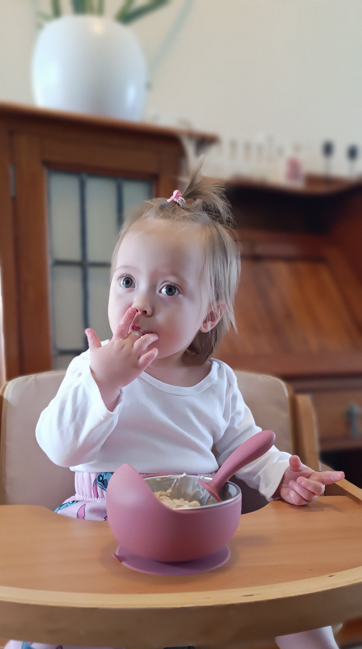 A toddler sitting in a wooden high chair at a dining table, eating from a pink silicone bowl with a pink spoon. The child is wearing a white long-sleeve shirt and has a small pink hair clip.