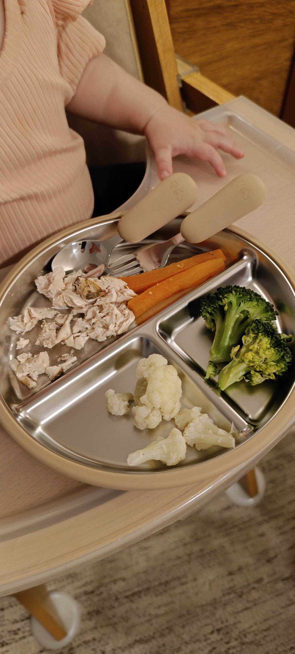 A close-up of a baby's hand resting on a beige high chair tray holding a stainless steel three-compartment meal plate with chicken, carrots, broccoli, and cauliflower. The baby is wearing a light pink ribbed top.