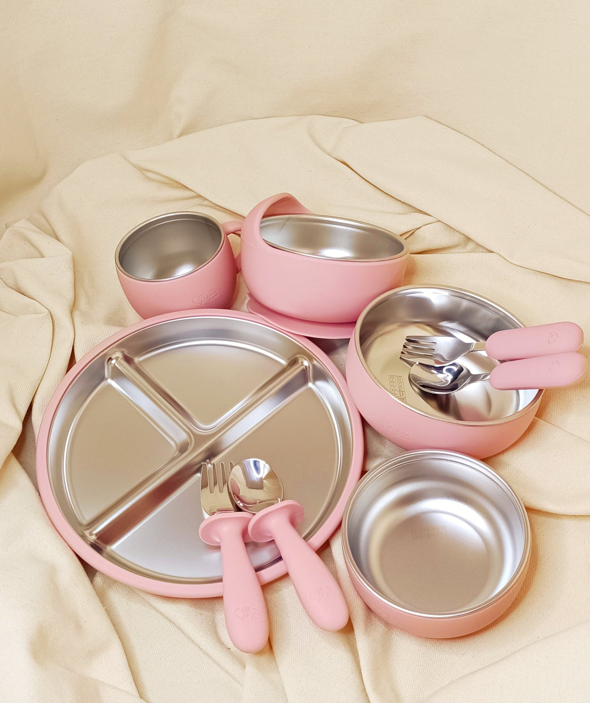 Pink children's silicone and stainless steel dinner set with plates, bowls, and utensils on a beige fabric background
