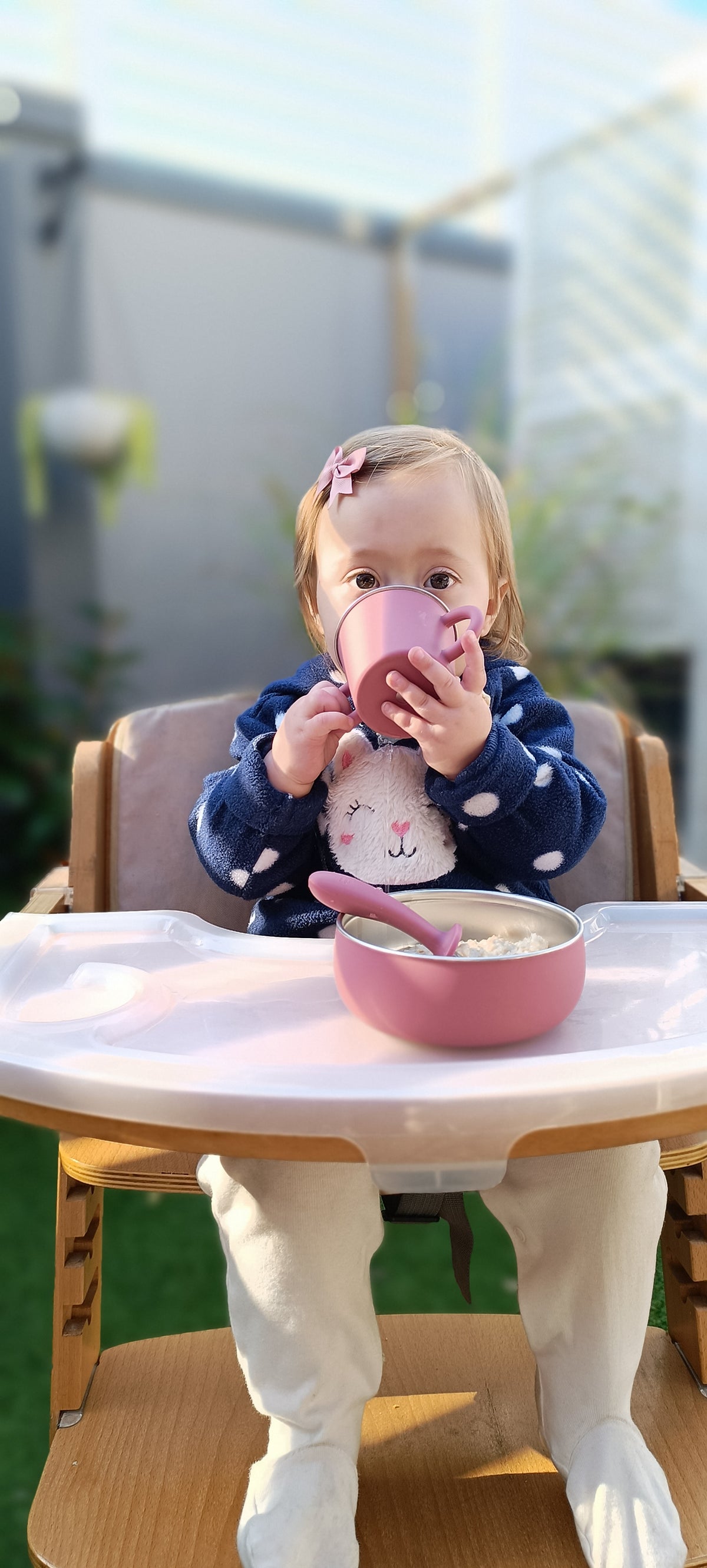 A toddler sitting in a wooden high chair outdoors, drinking from a pink cup with a matching pink spoon and a pink bowl of cereal on the tray. The child wears a navy blue polka-dot top and white pants.